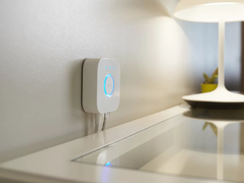 Philips Hue Bridge älyvalaistuksen ohjauskeskus valkoisella seinällä ja pöydällä lampun vieressä.